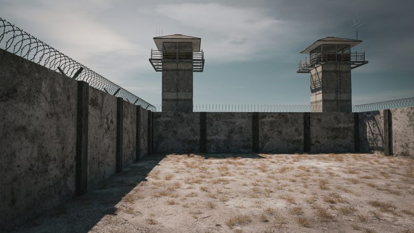 Concrete prison walls with two guard towers and a sandy courtyard under a cloudy sky.